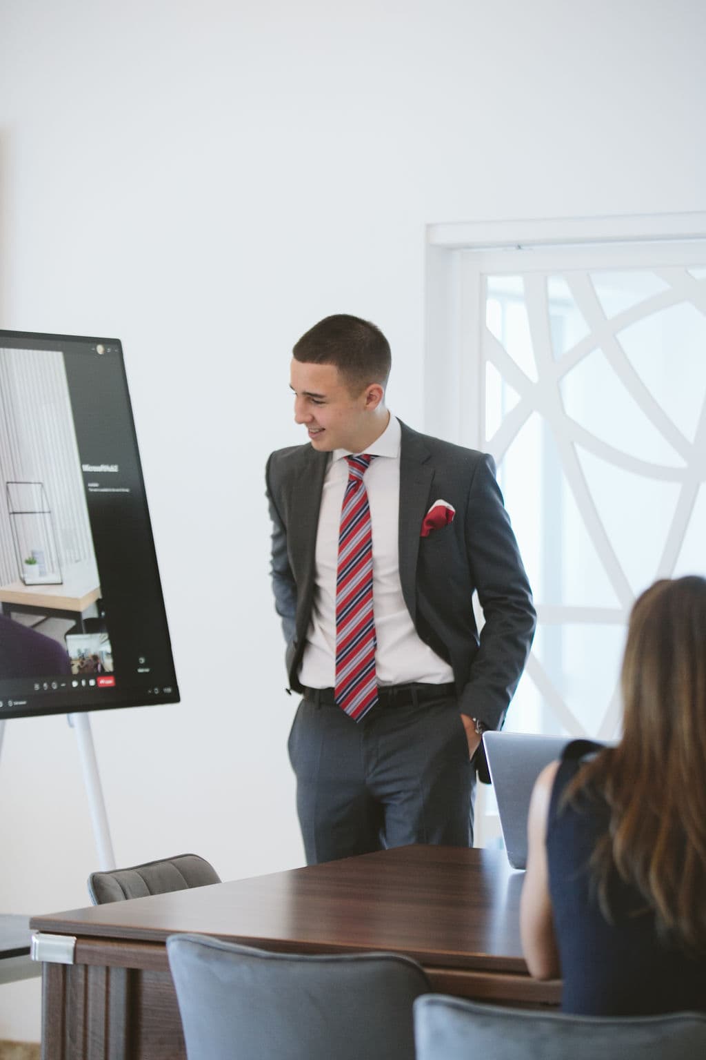 Smiling man in a suit stands by a digital screen during a professional office meeting.