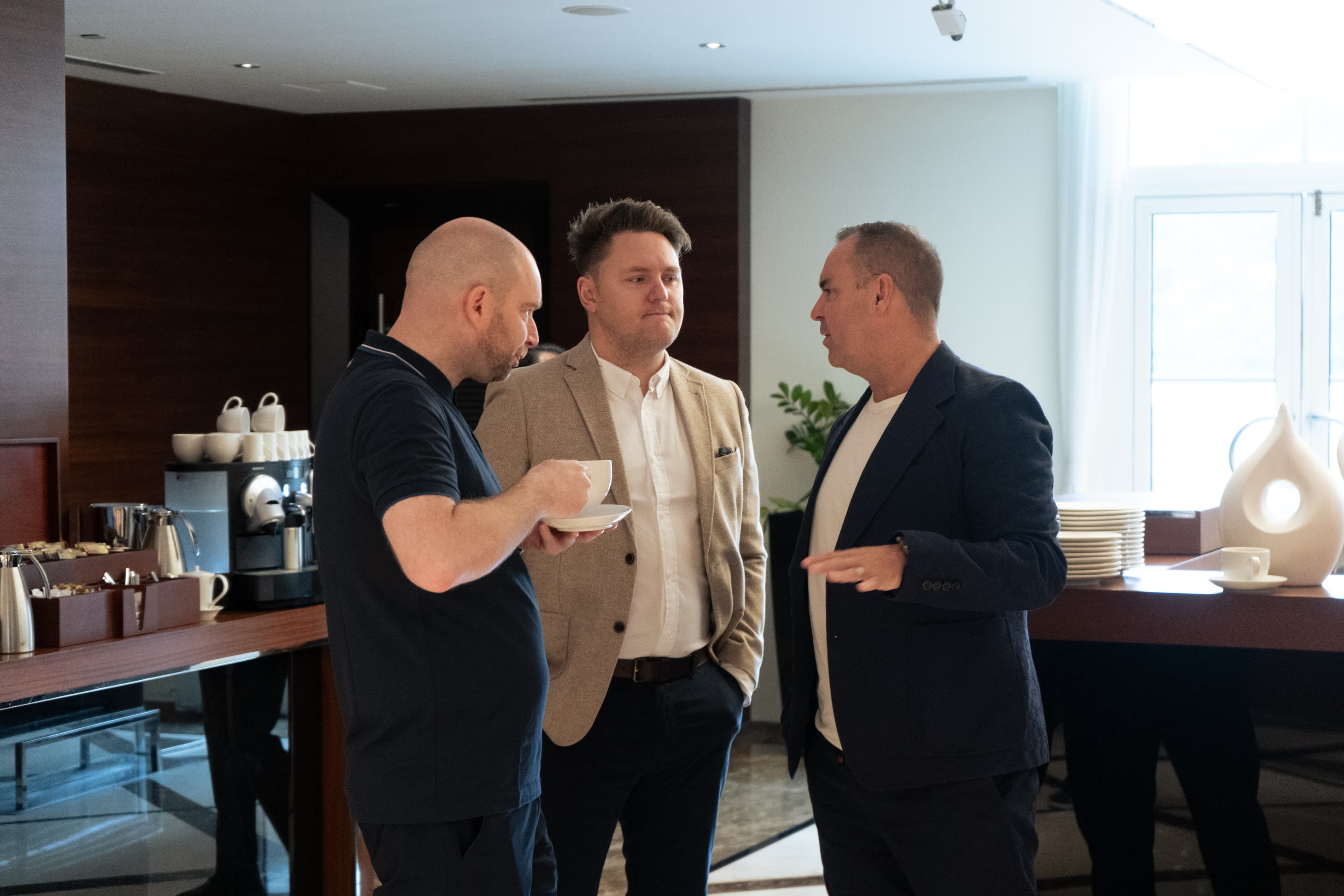 Three men in business casual attire talk near a coffee station in a modern lounge.