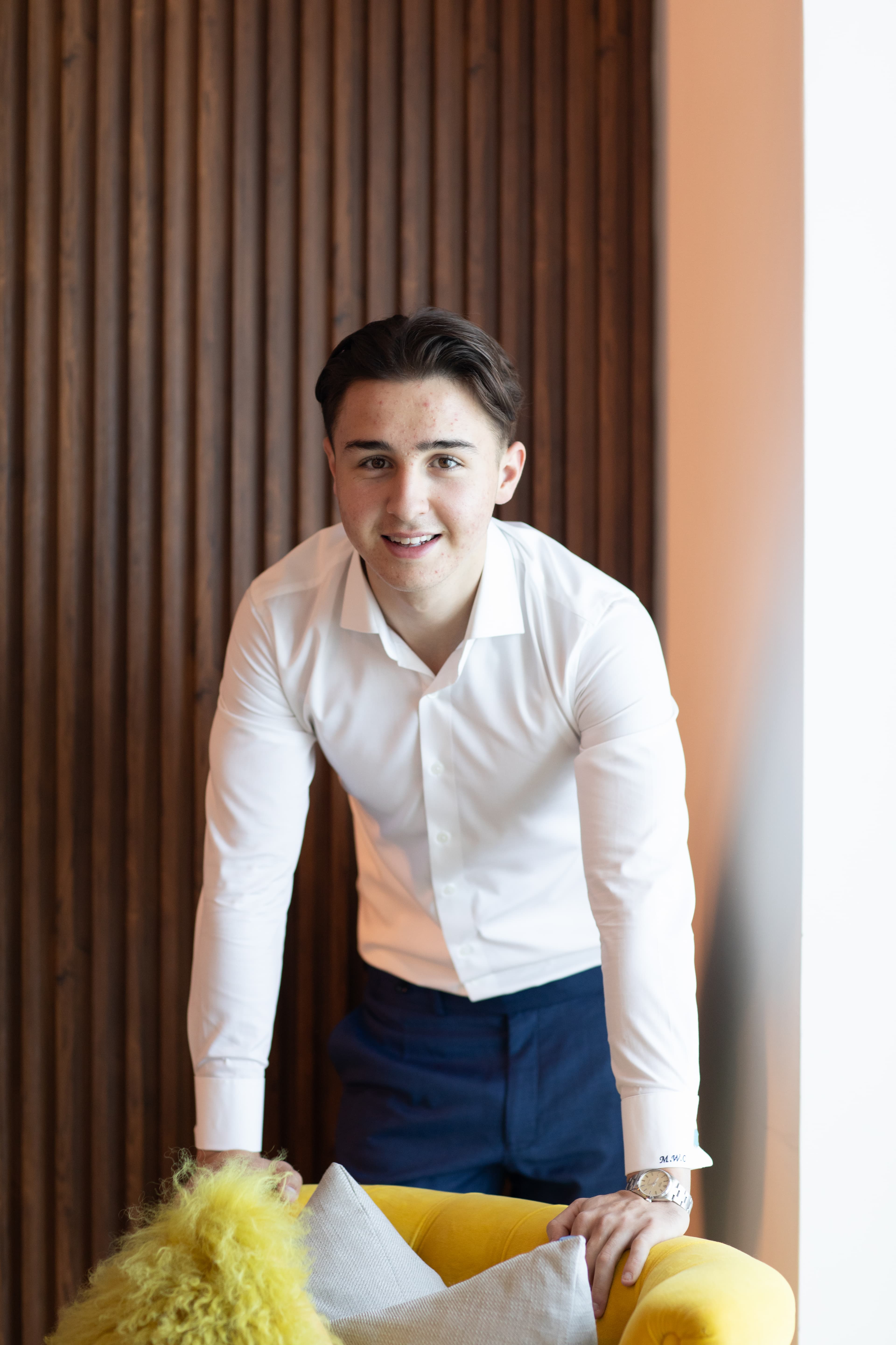 Smiling young man in a white shirt leans forward over a yellow velvet armchair.