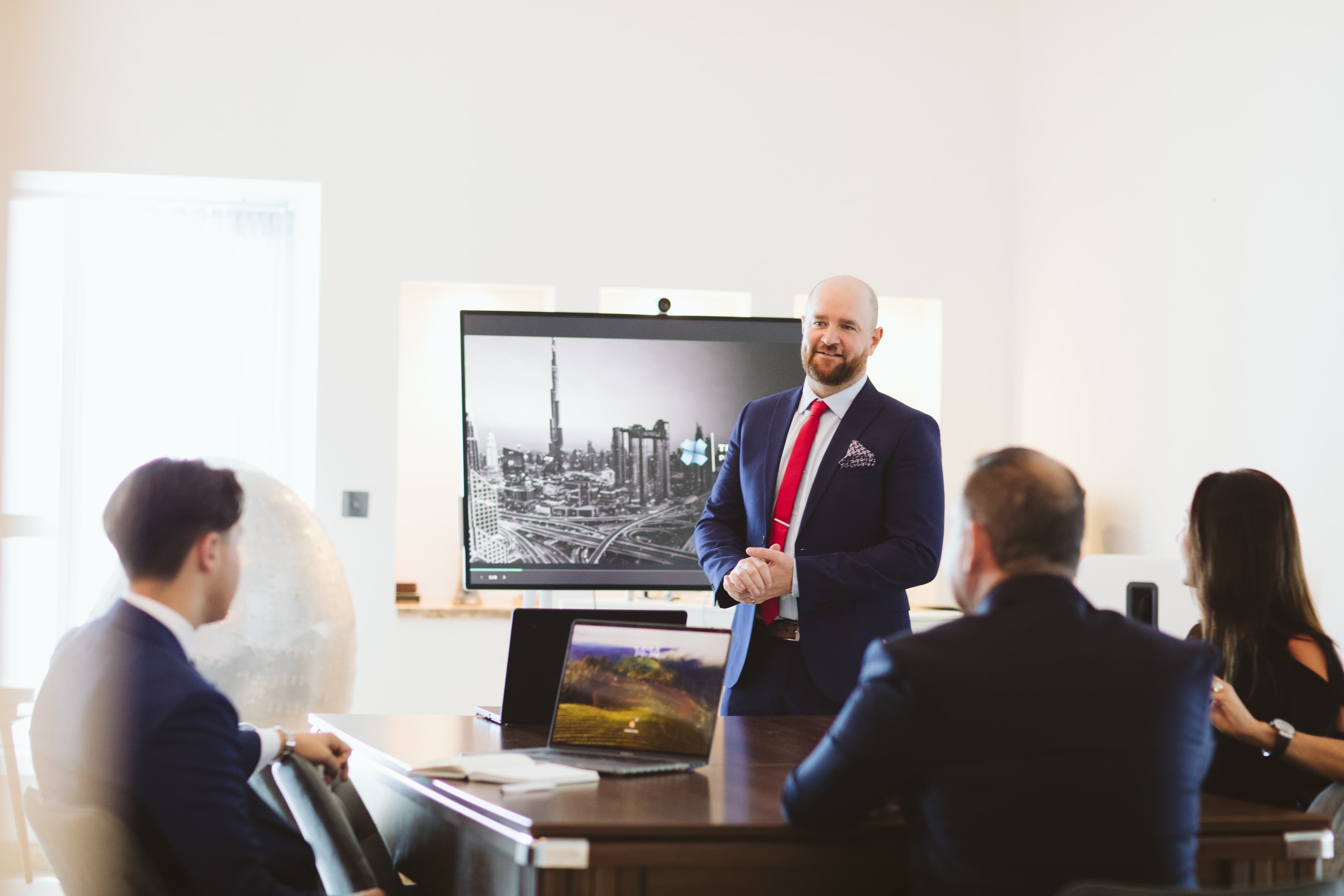 Businessman in a blue suit presenting to colleagues in a bright, modern office setting.