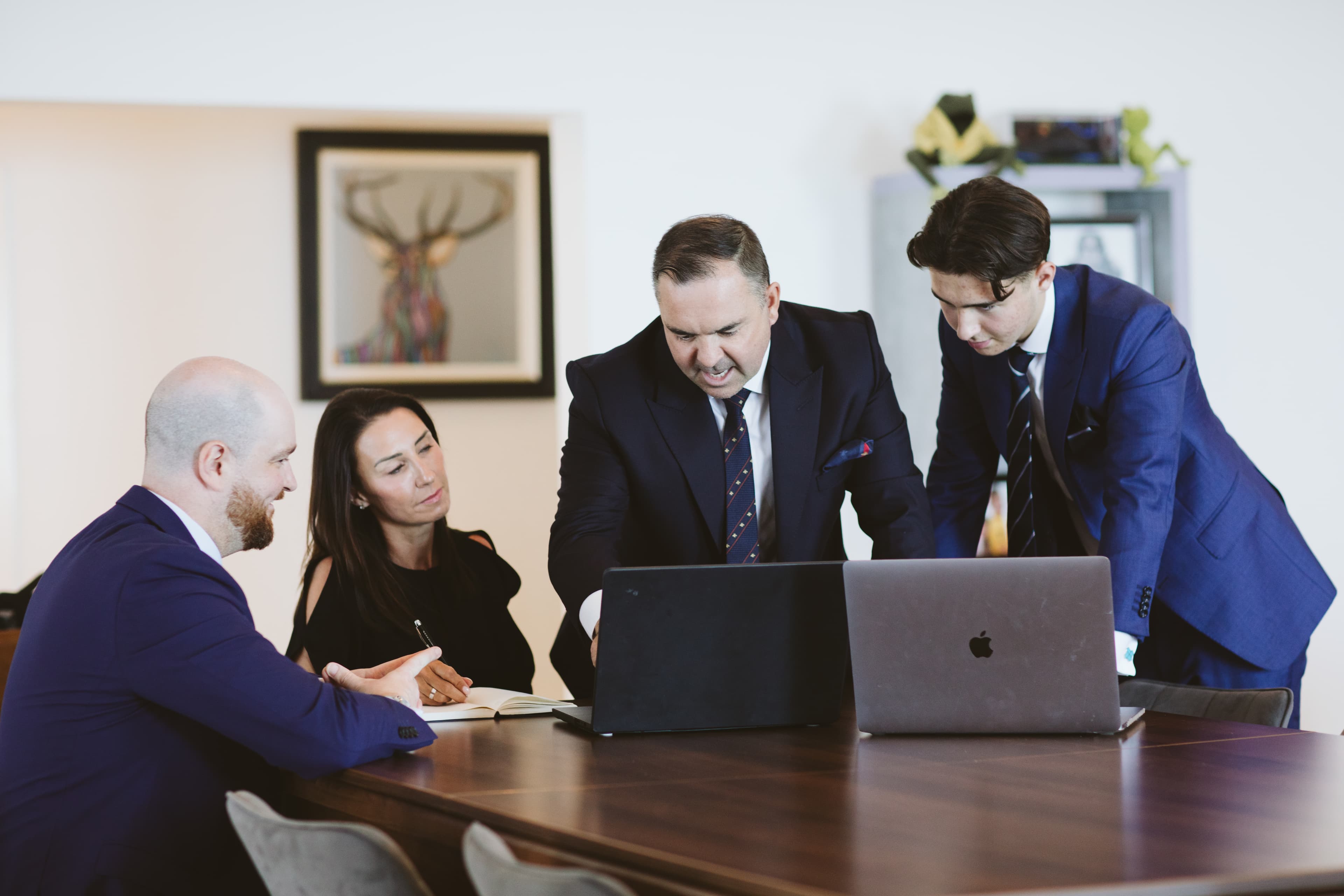 Four business professionals in suits collaborate around a table, focused on a laptop screen.