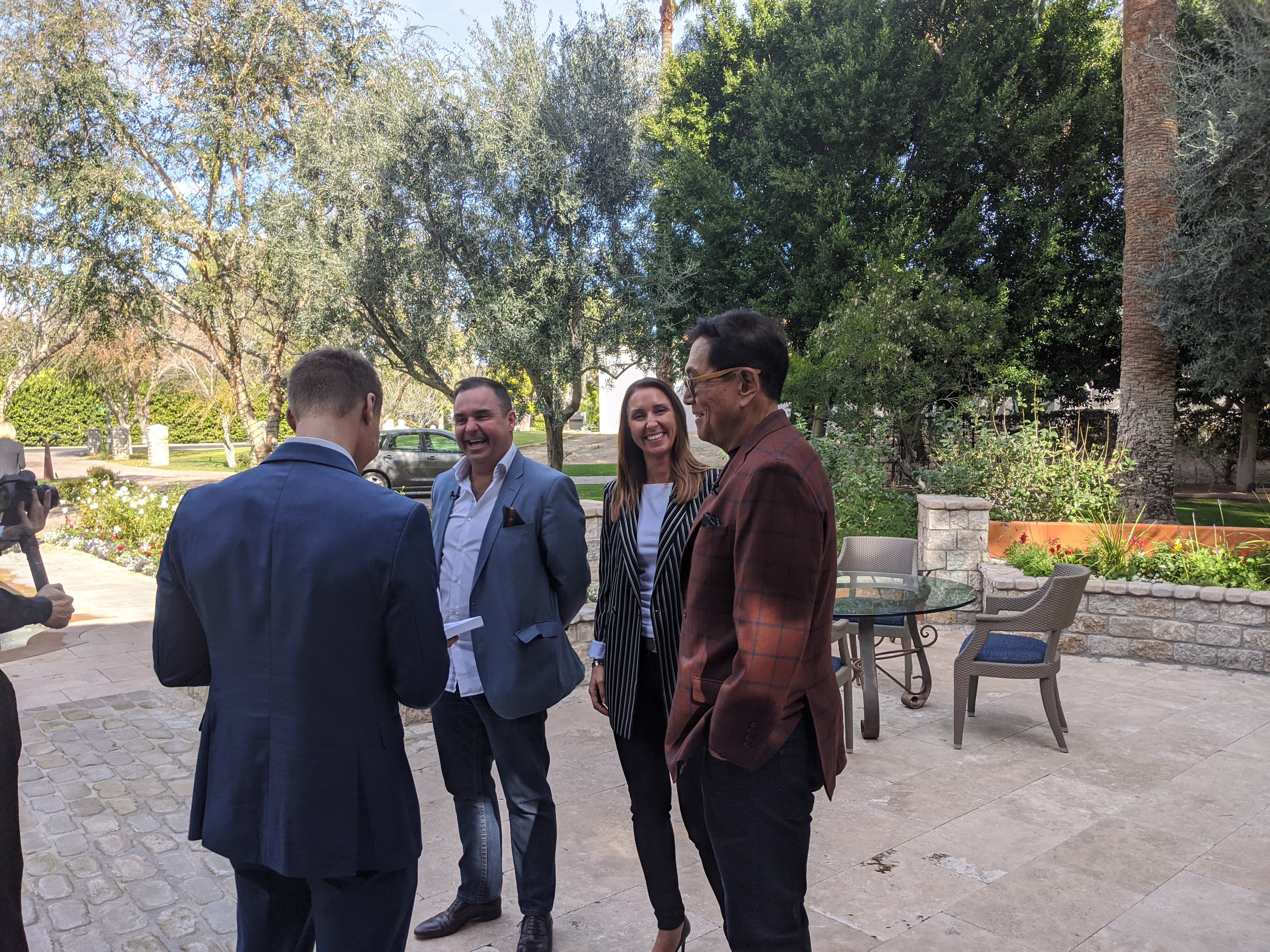 Four professionals in blazers stand on a stone patio, laughing and talking in a garden.